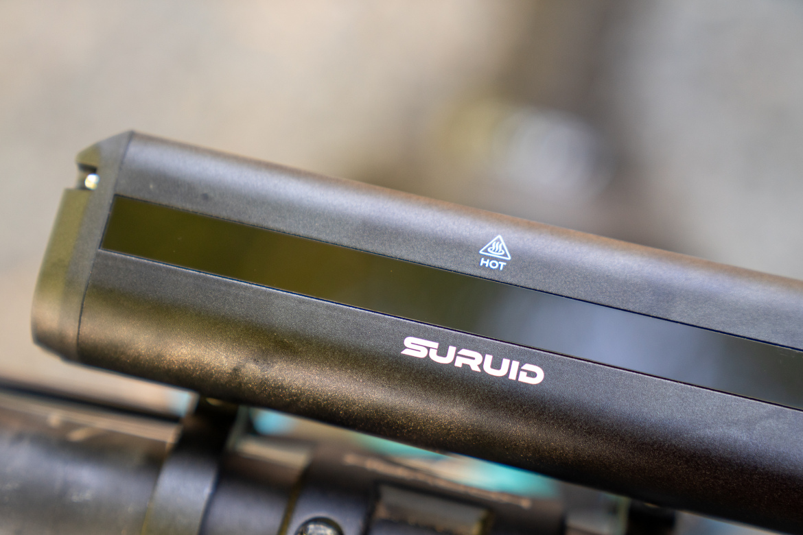 Close-up of a black bicycle light attachment with a warning symbol indicating "HOT" and the brand name "SURUID" displayed prominently. The light is mounted on a bicycle handlebar.