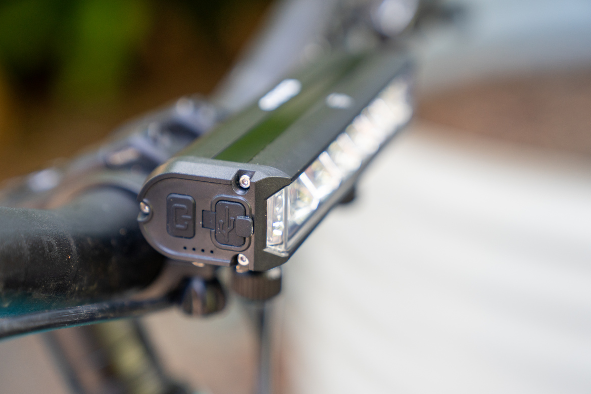 Close-up of a bicycle handlebar light mounted on the side, featuring multiple LED bulbs and buttons for power and charging. The background is blurred, emphasizing the light and its details.