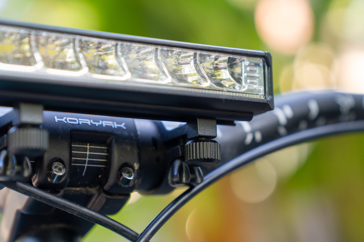 Close-up of a bicycle handlebar with a mounted LED light, showing the brand name "KORYAK" and details of the light's attachment mechanism and the handlebar itself. The background features a blurred, natural setting with soft bokeh.