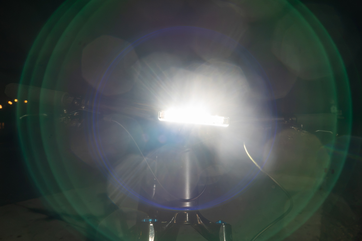 A bright bicycle headlight illuminating the darkness, creating a lens flare effect with circular light patterns in the foreground.