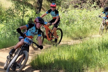 A group of young cyclists riding mountain bikes along a dirt trail, surrounded by greenery and trees. One cyclist is leaning into a turn, while two others are visible in the background. All riders are wearing helmets and matching team jerseys.