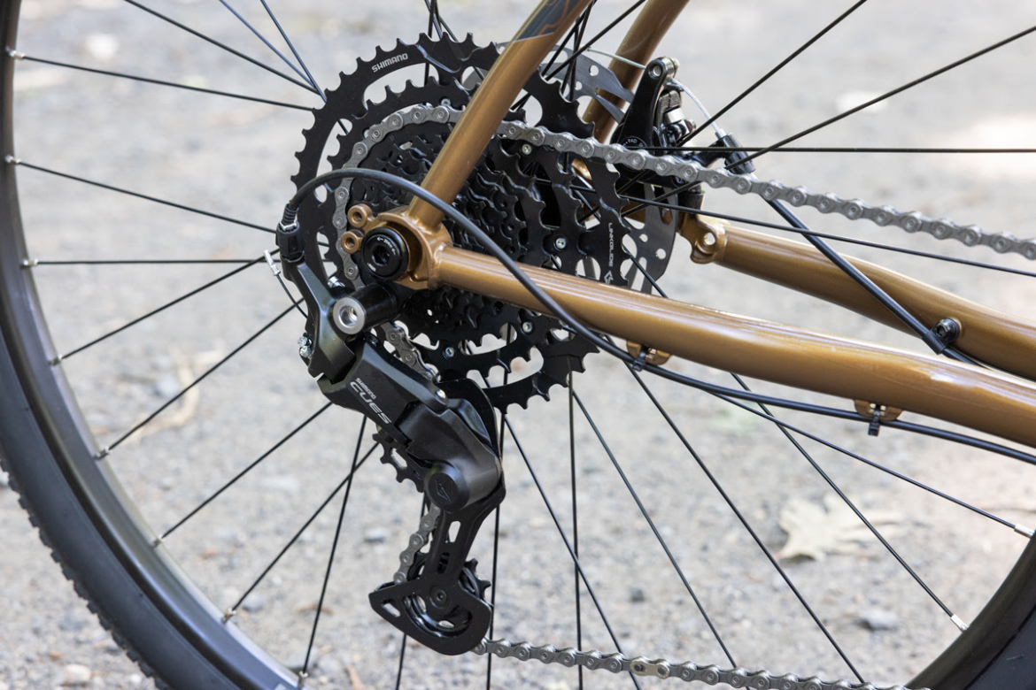 Close-up view of the rear derailleur and cassette of a bicycle, highlighting details such as the chain, gears, and metallic components against a gravel background.