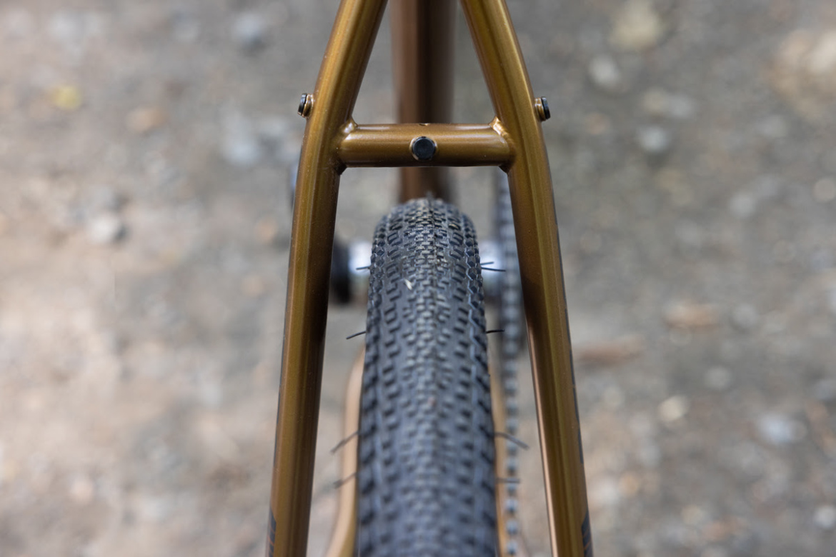 Close-up view of a bicycle tire seen through the frame’s rear triangle, showcasing the textured tread and gold-colored frame against a blurred gravel background.