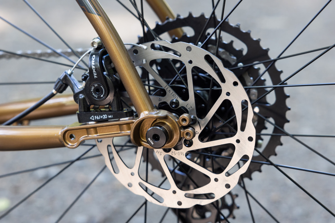 Close-up image of a bicycle's rear braking system, featuring a metallic disc brake rotor and caliper, with a focus on the intricate design of the rotor and the attachment to the bike frame. The background shows a blurred ground surface, highlighting the bicycle components.
