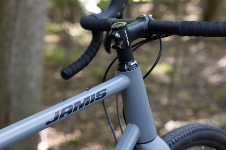 Close-up view of a grey bicycle frame featuring the brand name "JAMIS" prominently displayed. The handlebars, with black grips, and the headset are visible, set against a blurred background of trees and foliage, suggesting an outdoor setting.