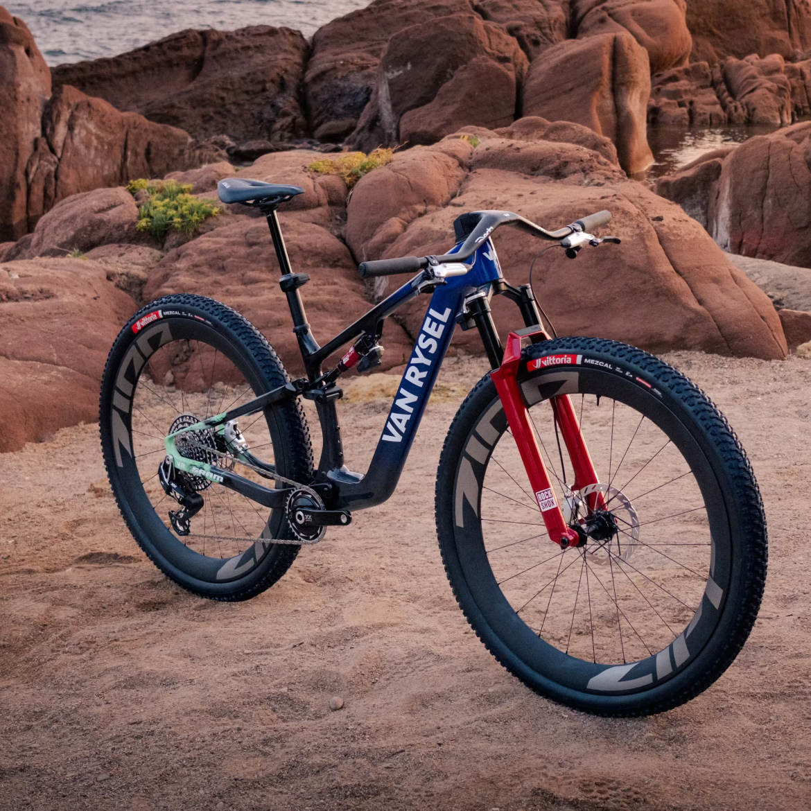 A mountain bike with a black and blue frame featuring the brand name "VAN RYSEL" prominently displayed. The bike has red front forks and thick, rugged tires, positioned on sandy ground near large, reddish-brown rocks and a body of water in the background. The scene suggests a picturesque outdoor setting ideal for cycling.