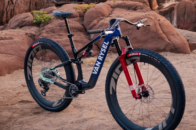 A mountain bike with a black and blue frame featuring the brand name "VAN RYSEL" prominently displayed. The bike has red front forks and thick, rugged tires, positioned on sandy ground near large, reddish-brown rocks and a body of water in the background. The scene suggests a picturesque outdoor setting ideal for cycling.