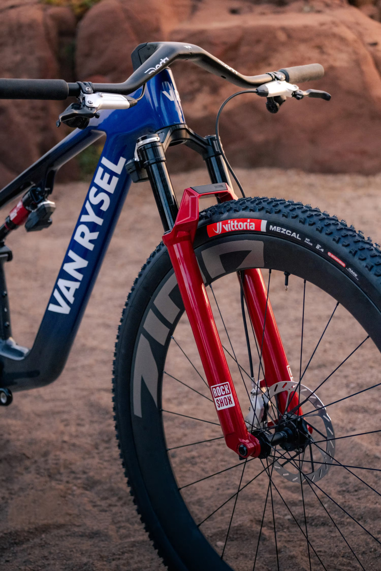 Close-up of a mountain bike featuring a blue frame with the text "VAN RYSEL," a red front fork labeled "ROCK SHOX," and a black tire with the brand "Vittoria Mezcal." The background shows a natural landscape with rocky formations.