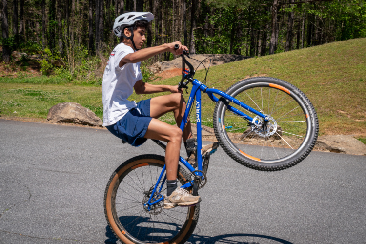 A young person wearing a helmet performs a wheelie on a blue bicycle on a paved road, surrounded by green grass and trees in the background.